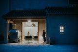 A person stands in the doorway of a rustic building at night, with a bright interior visible inside and a dark exterior lit by blue ambient light. A barrel, a covered appliance, and a small window are visible on the right side of the structure.