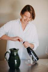 A woman in a white button-down shirt smiles while opening a dark bottle, with a dark green pitcher and a wine glass on a table in a softly lit indoor setting.